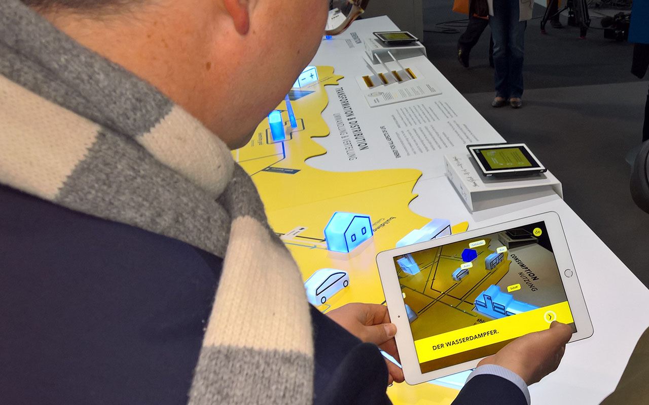 Ein Besucher hält ein Tablet über ein Ausstellungsmodell; auf dem Bildschirm erscheinen zusätzliche 3D-Objekte und Beschriftungen