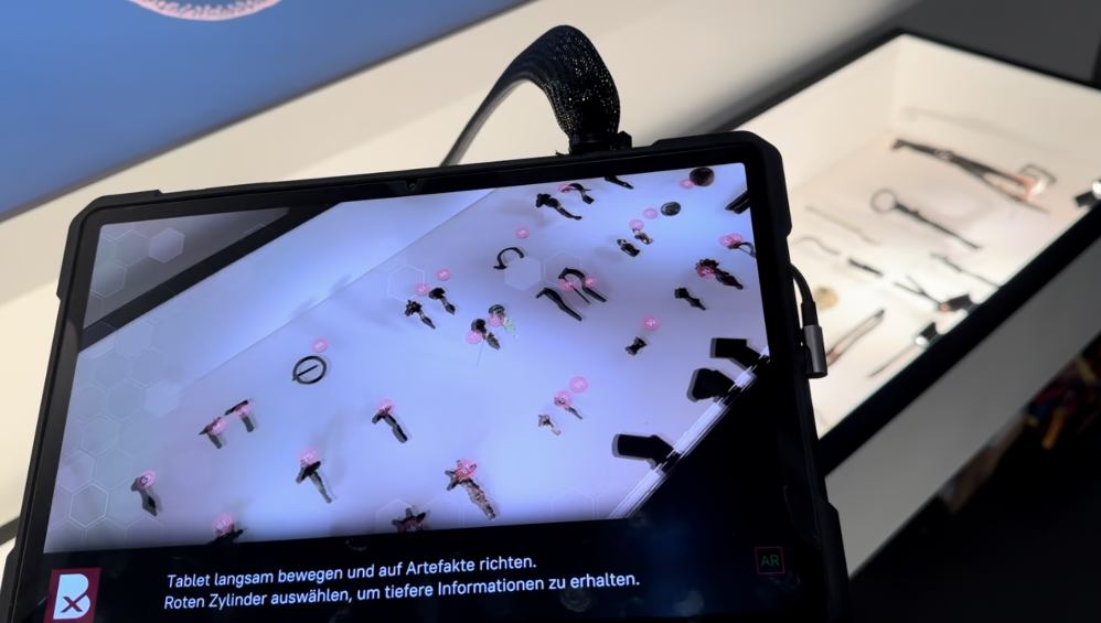 Tablet mit AR-App vor einer Vitrine, eingeblendete Markierungen über den Grabfunden